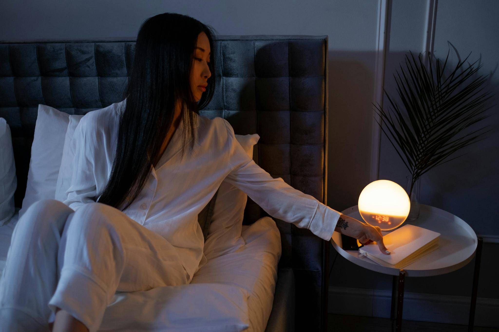 Asian woman in pajamas adjusting a glowing spherical lamp on a nightstand in a dimly lit bedroom.
